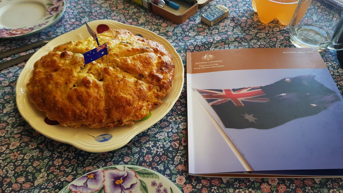 A large scone with a small Australian flag on a floral tablecloth, beside an Australian Government booklet — a country Queensland table.