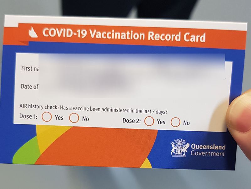 A Queensland Government COVID-19 vaccination record card, held while waiting in the queue. The name and date of birth are blurred.